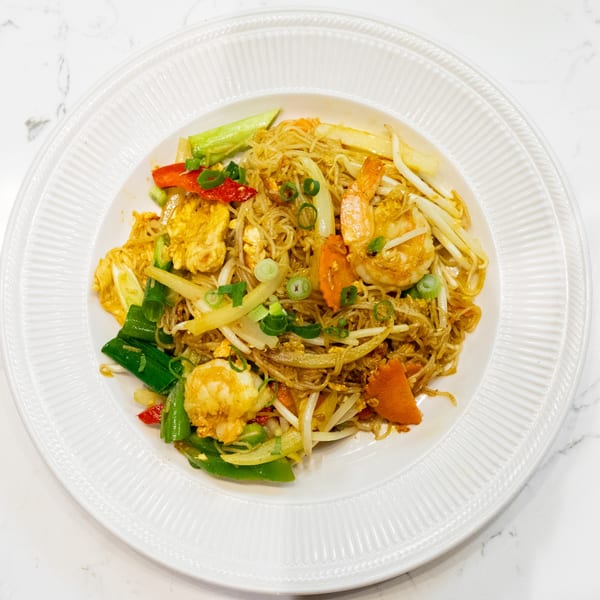A white plate filled with Singapore noodles, shrimp, chicken, and vegetables on a marble surface.