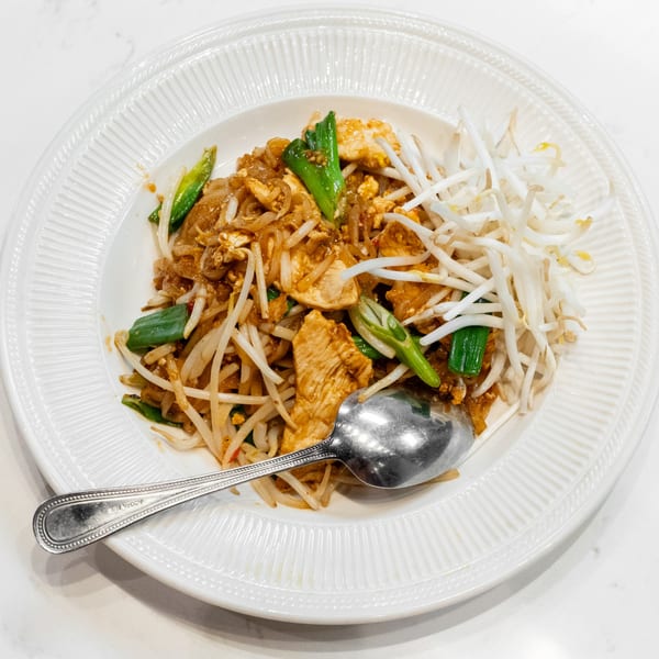 A white plate filled with chicken pad thai, garnished with green onions and bean sprouts, with a spoon.