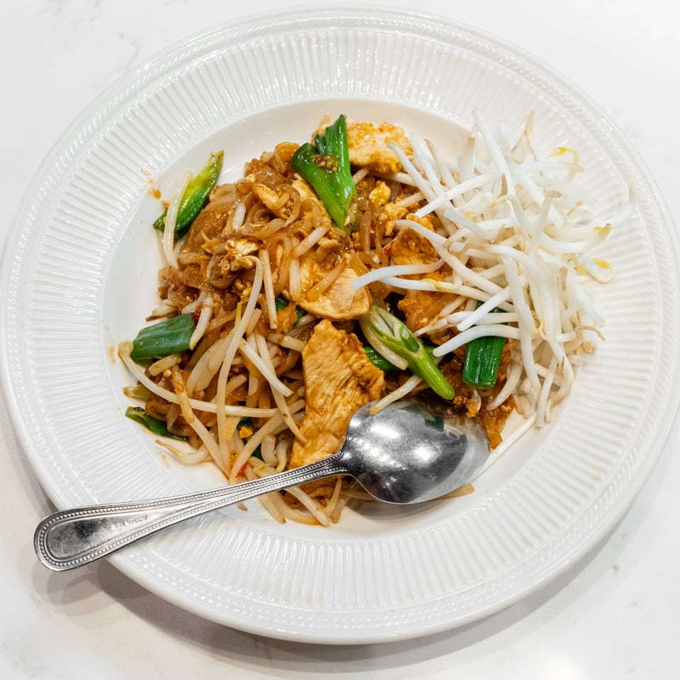 A white plate filled with chicken pad thai, garnished with green onions and bean sprouts, with a spoon.