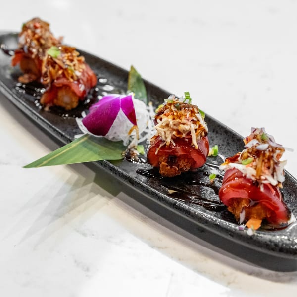 Four pieces of tuna tataki, drizzled with sauce and topped with shredded vegetables, on a black plate.