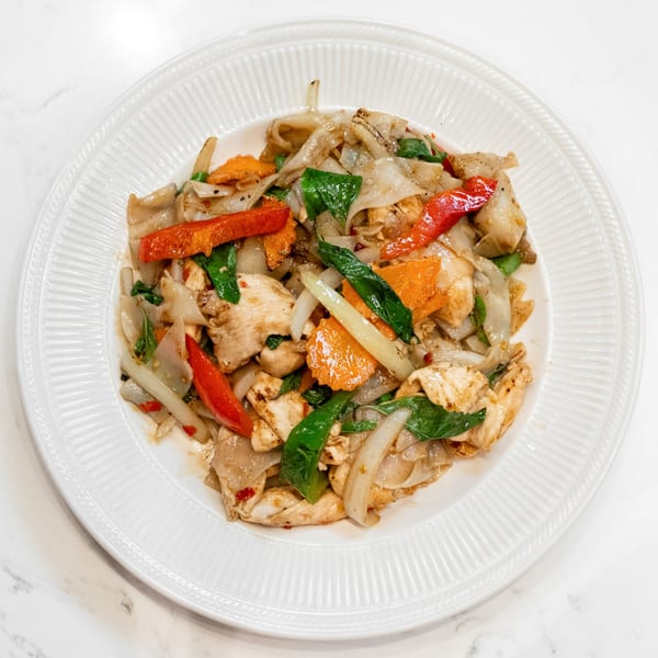 Pad See Ew with chicken, wide noodles, red bell peppers, carrots, and green basil on a white plate.